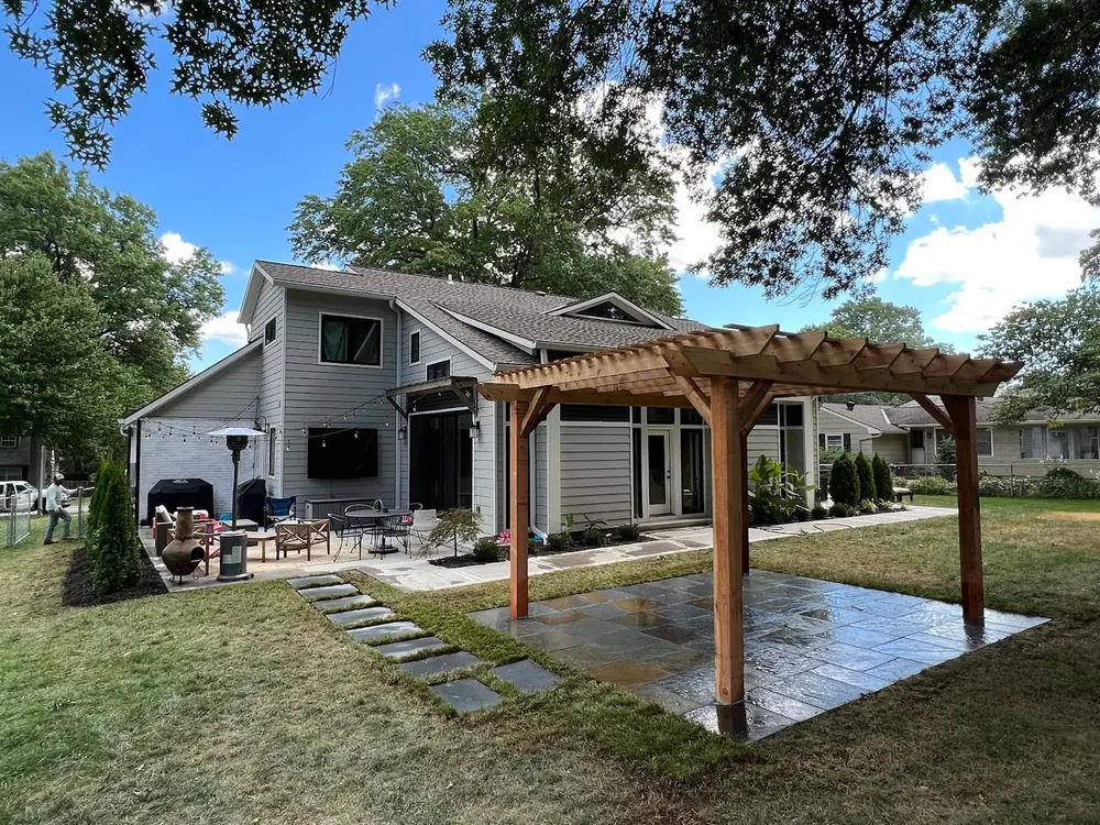 Cedar pergola over a paver patio with a green lawn and mature trees behind a home