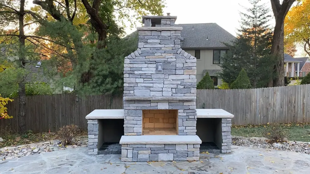 Stone outdoor fireplace with a tall chimney and flagstone hearth in a fenced backyard